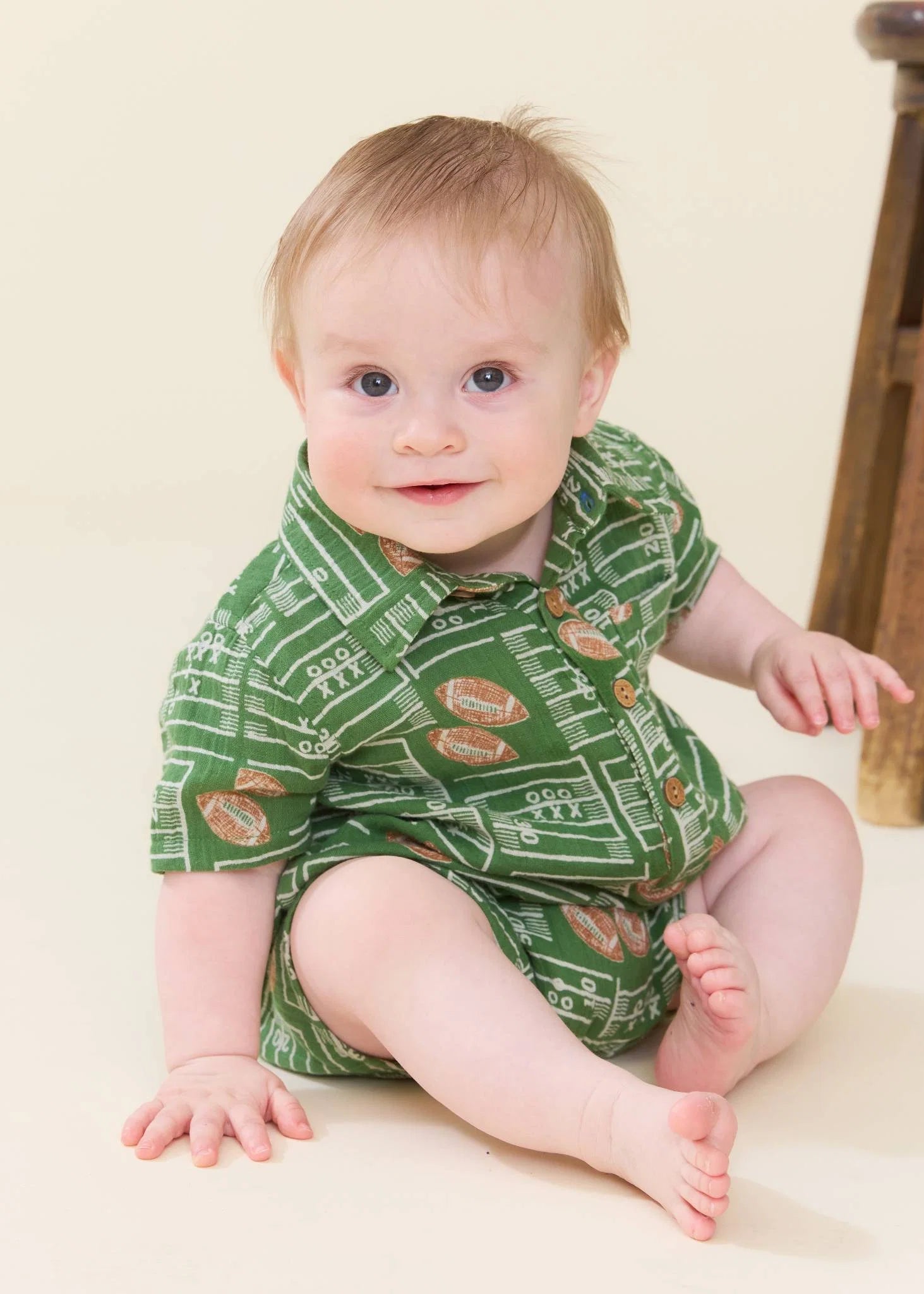 A baby sitting on a cream-colored floor wearing a green short-sleeve romper with a football field and football pattern, next to a wooden stool.