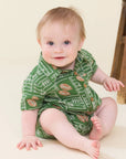 A baby sitting on a cream-colored floor wearing a green short-sleeve romper with a football field and football pattern, next to a wooden stool.