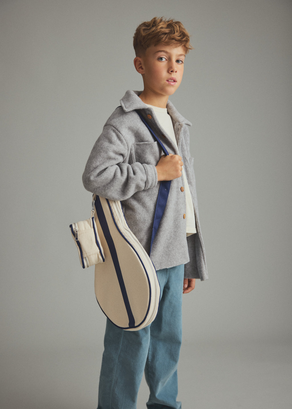 A young boy wearing a gray button-up shirt jacket over a white t-shirt and blue jeans, carrying a cream canvas tennis bag with navy blue stripes and a matching small pouch against a solid gray studio background.