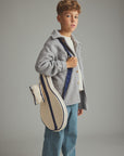 A young boy wearing a gray button-up shirt jacket over a white t-shirt and blue jeans, carrying a cream canvas tennis bag with navy blue stripes and a matching small pouch against a solid gray studio background.