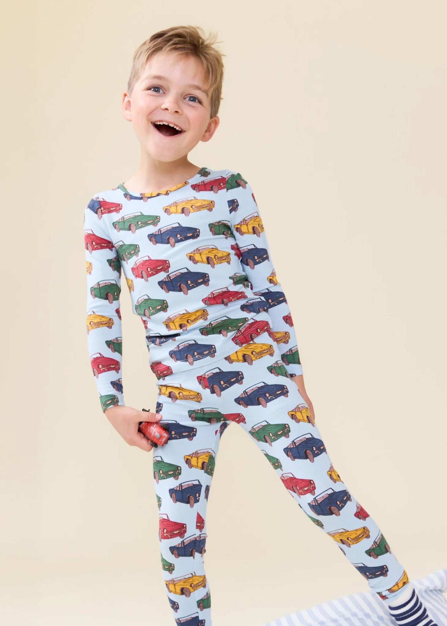A young boy smiling and wearing a light blue long-sleeve pajama set with an all-over colorful car print, standing against a plain beige background.