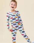 A young boy smiling and wearing a light blue long-sleeve pajama set with an all-over colorful car print, standing against a plain beige background.
