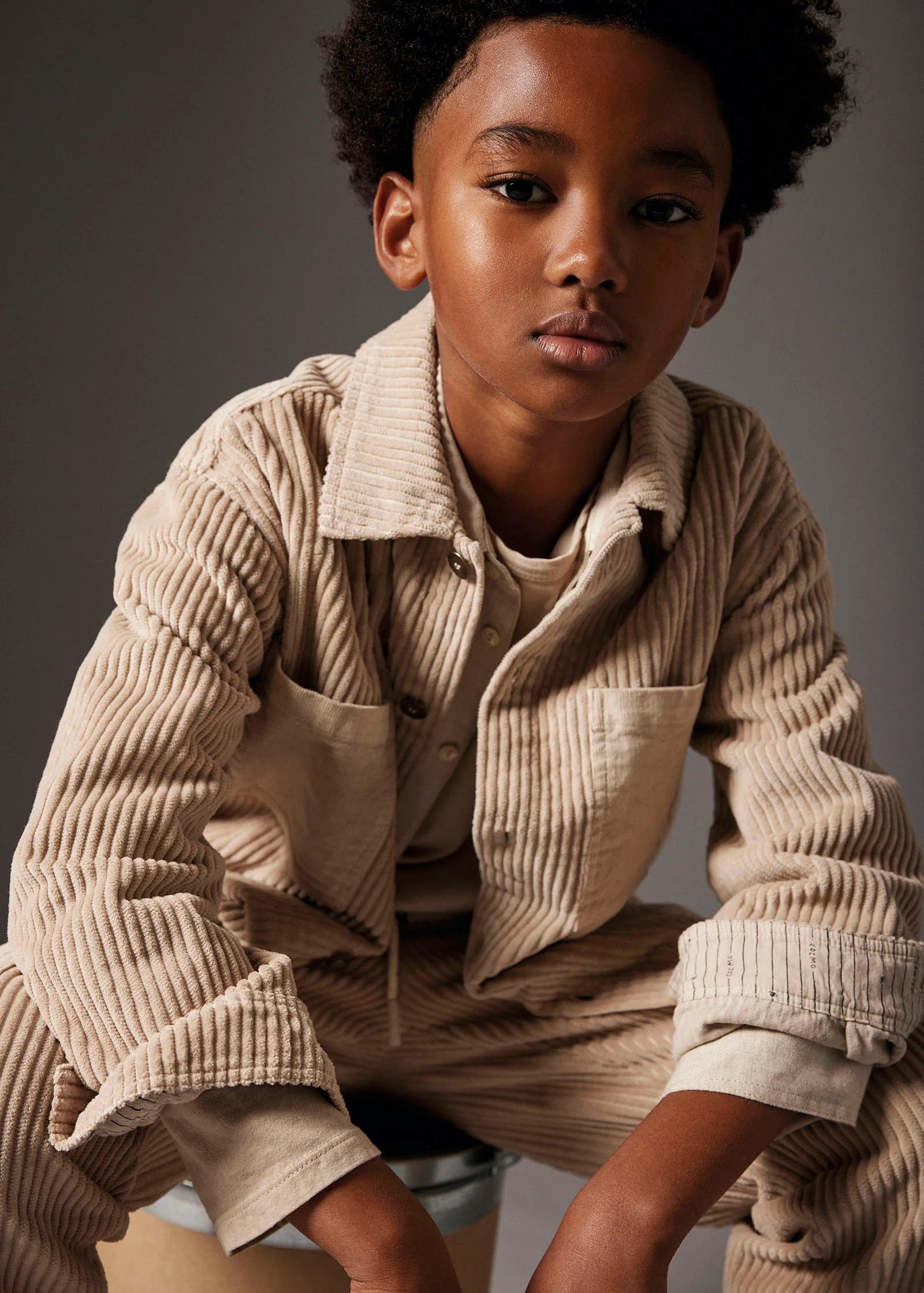 A young boy wearing a beige corduroy button-up shirt and matching corduroy pants, posed against a dark gray studio background.