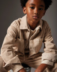 A young boy wearing a beige corduroy button-up shirt and matching corduroy pants, posed against a dark gray studio background.