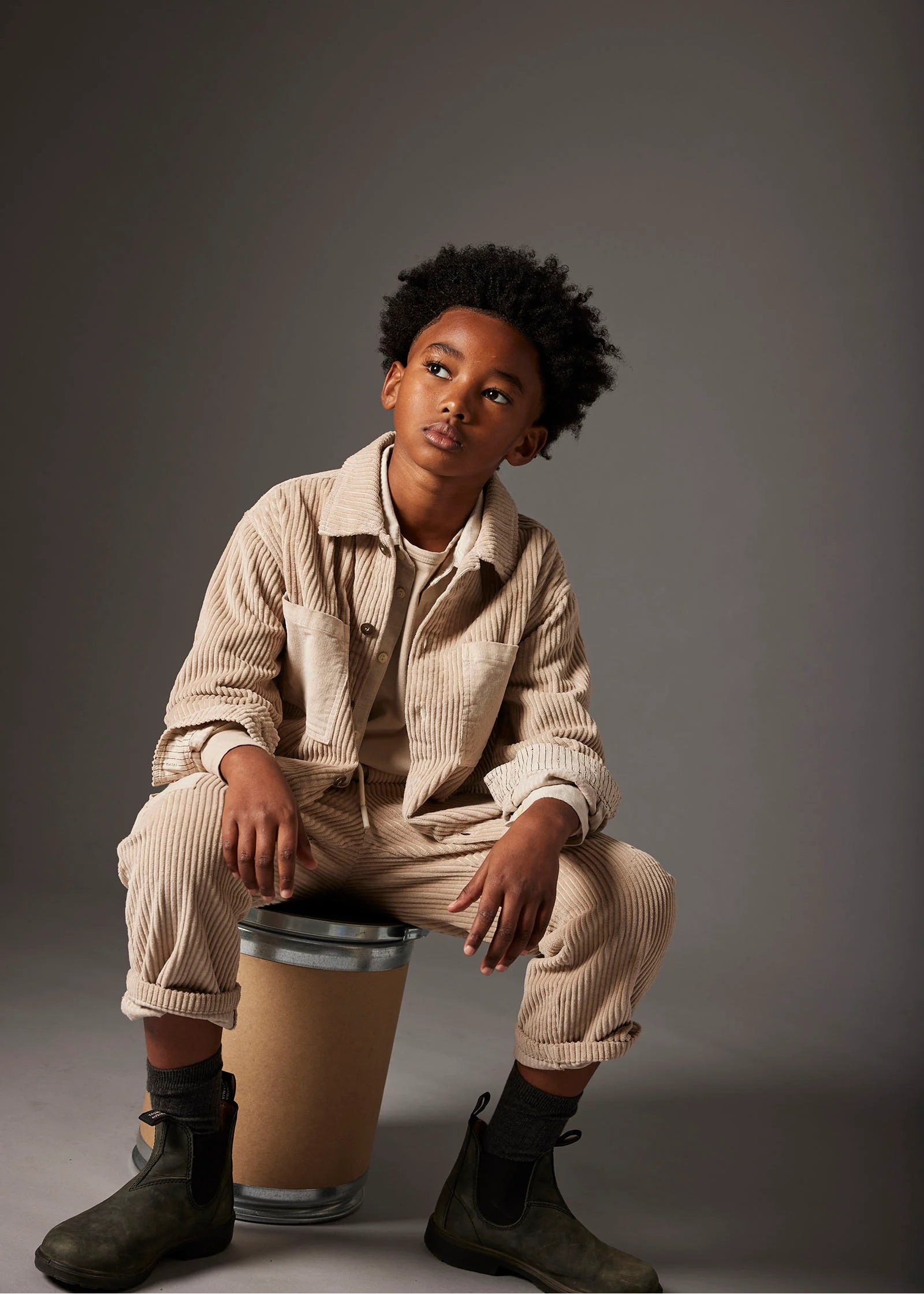 A young boy with curly black hair sits on a cardboard shipping box, wearing a beige corduroy button-up shirt and matching corduroy pants, dark gray socks, and dark boots against a dark gray studio background.