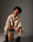 A young boy with curly black hair sits on a cardboard shipping box, wearing a beige corduroy button-up shirt and matching corduroy pants, dark gray socks, and dark boots against a dark gray studio background.