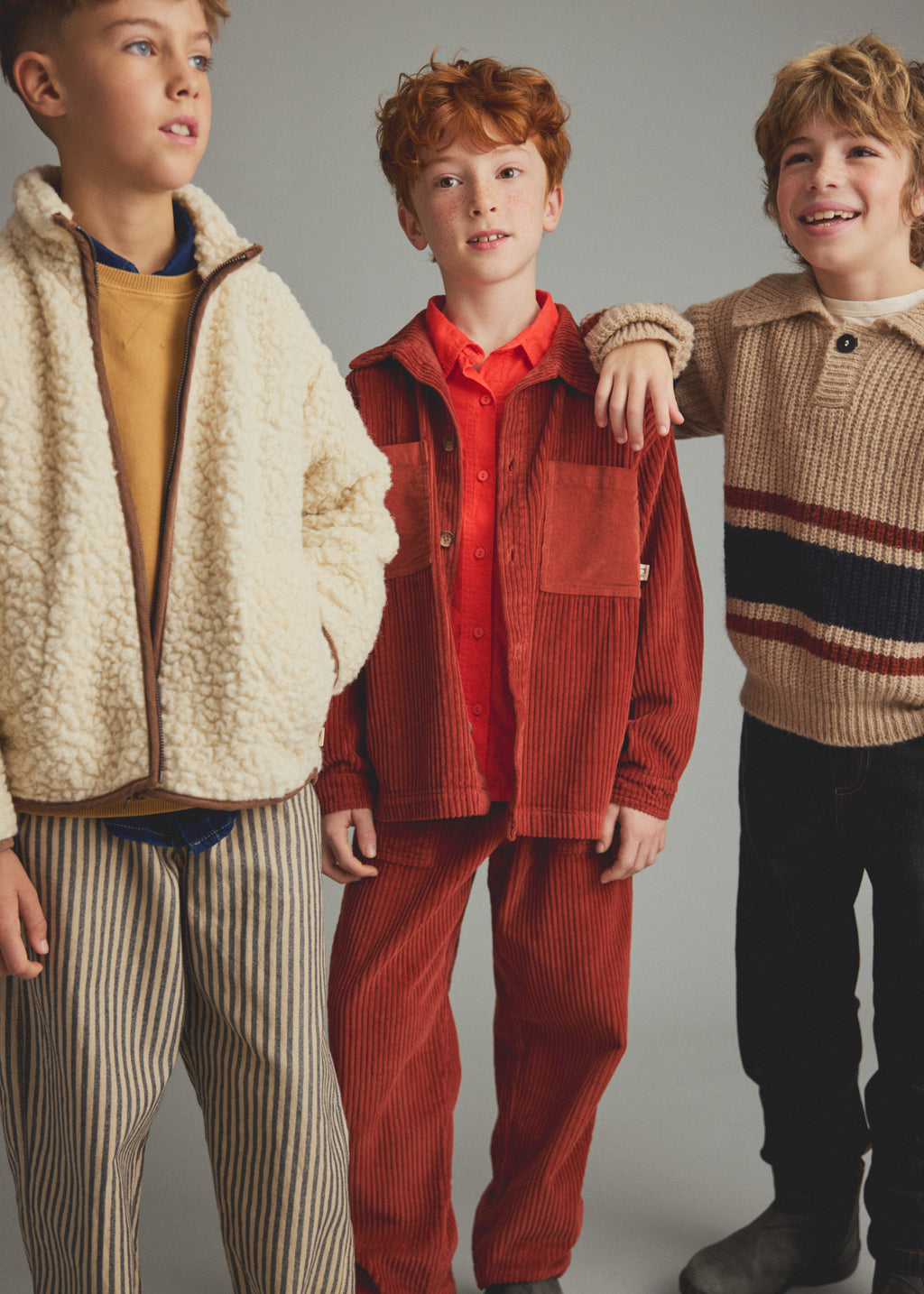 Three boys modeling children's clothing against a plain gray studio background. The boy on the left wears a cream sherpa zip jacket over a mustard top and striped pants.