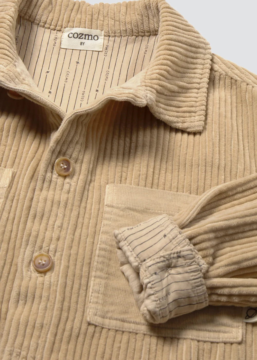 Close-up of a beige corduroy button-up shirt with a striped inner collar lining and a chest pocket, shown on a light neutral background.