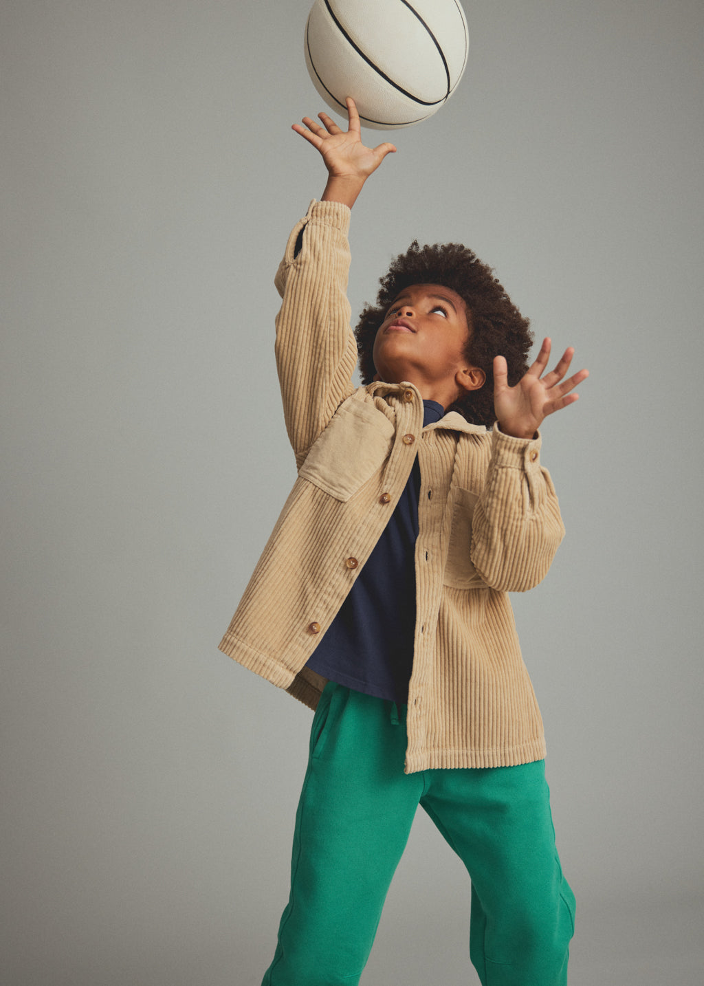 A child wearing a beige corduroy button-up jacket, a navy blue t-shirt, and green sweatpants reaches up toward a basketball against a solid gray studio background.