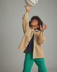 A child wearing a beige corduroy button-up jacket, a navy blue t-shirt, and green sweatpants reaches up toward a basketball against a solid gray studio background.