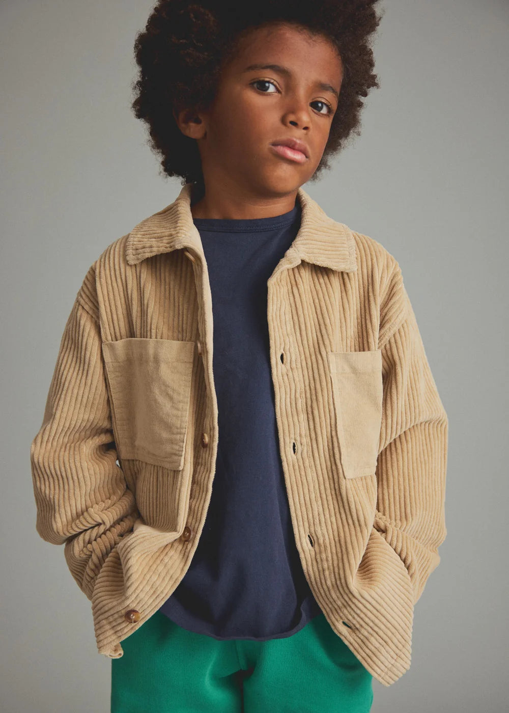 Child wearing a beige corduroy jacket over a navy shirt with green pants on a gray background
