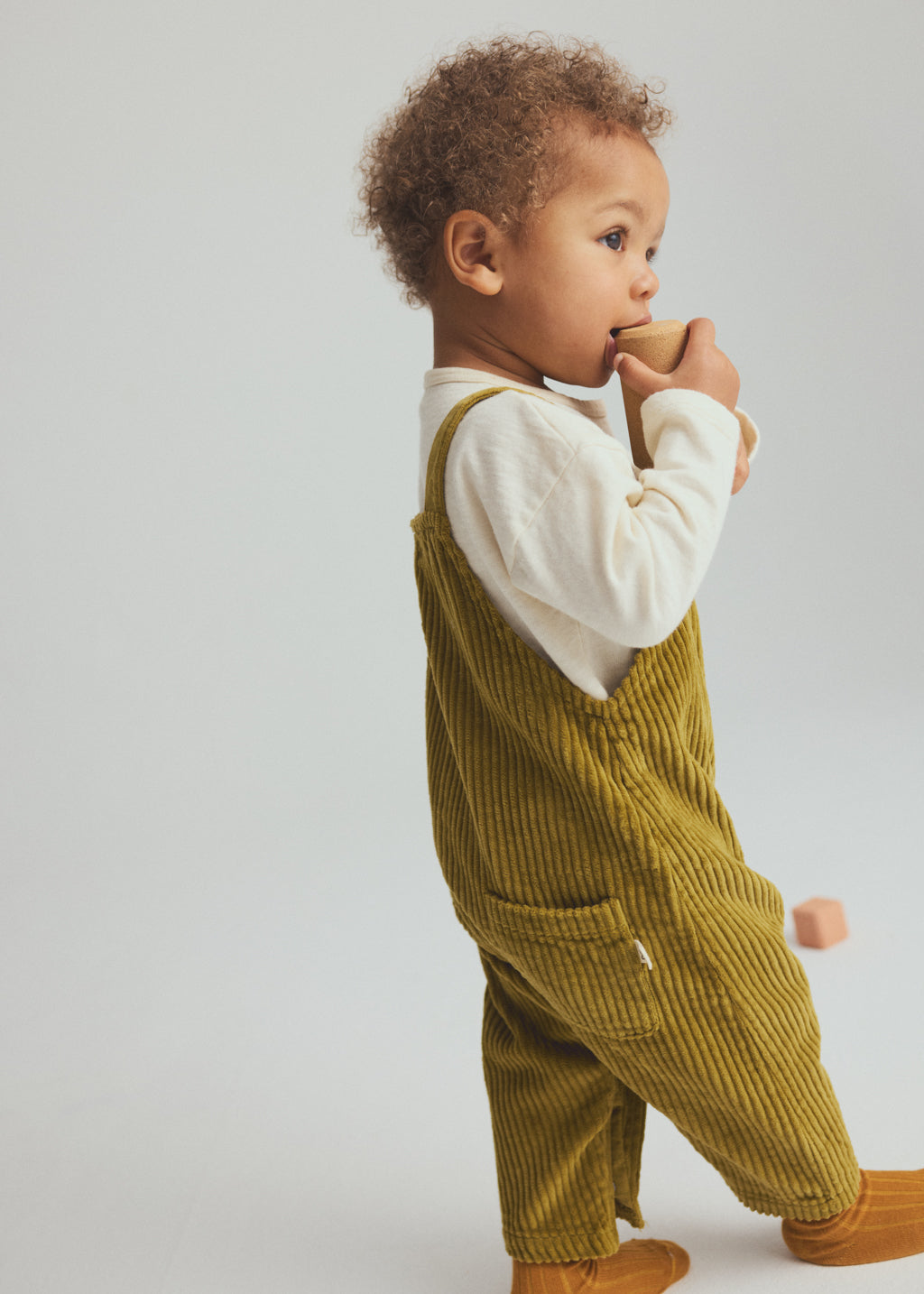 A toddler standing in profile wearing olive green corduroy overalls over a white long-sleeve shirt, with mustard yellow socks, against a light gray studio background.