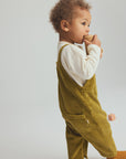 A toddler standing in profile wearing olive green corduroy overalls over a white long-sleeve shirt, with mustard yellow socks, against a light gray studio background.