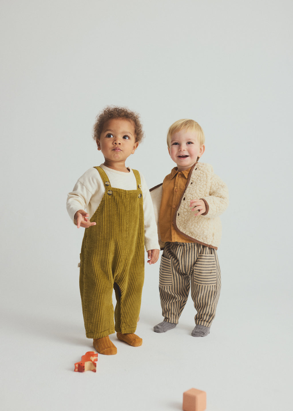 Two toddlers modeling coordinated autumn outfits in a studio setting. The child on the left wears olive corduroy overalls over a cream shirt, and the child on the right wears a cream textured cardigan over a mustard top with brown-and-beige striped pants.