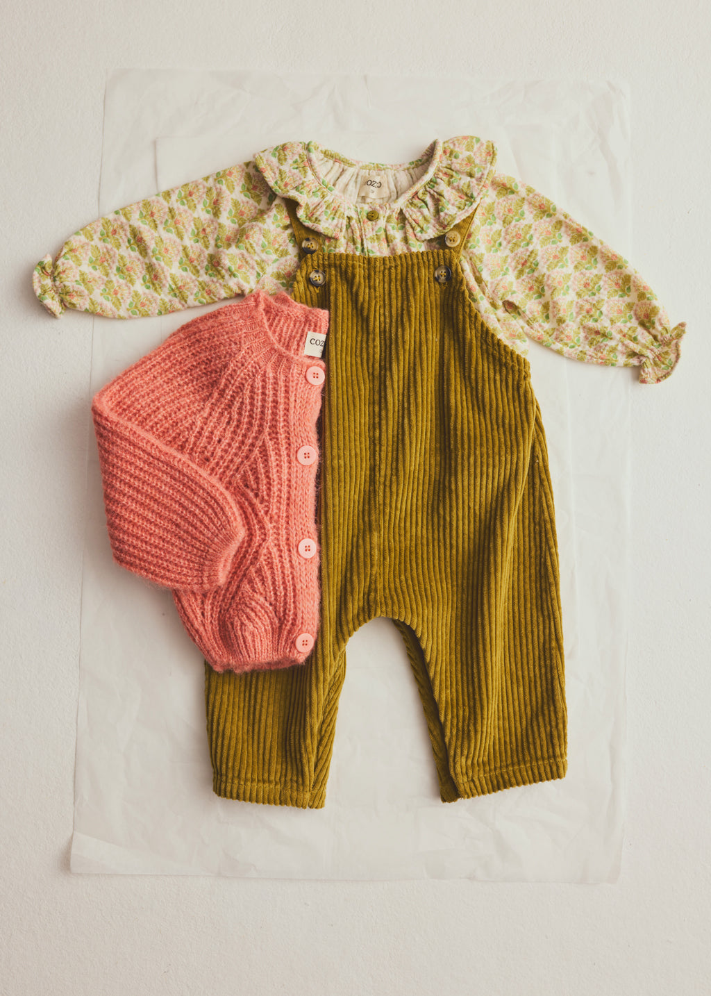 A baby clothing set laid flat on a white sheet, featuring olive green corduroy overalls, a pale yellow patterned long-sleeve top with a ruffled collar, and a coral pink knitted button-front cardigan.