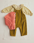 A baby clothing set laid flat on a white sheet, featuring olive green corduroy overalls, a pale yellow patterned long-sleeve top with a ruffled collar, and a coral pink knitted button-front cardigan.