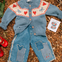 Children's blue knitted cardigan and jeans with heart and bow pattern on a wooden surface.