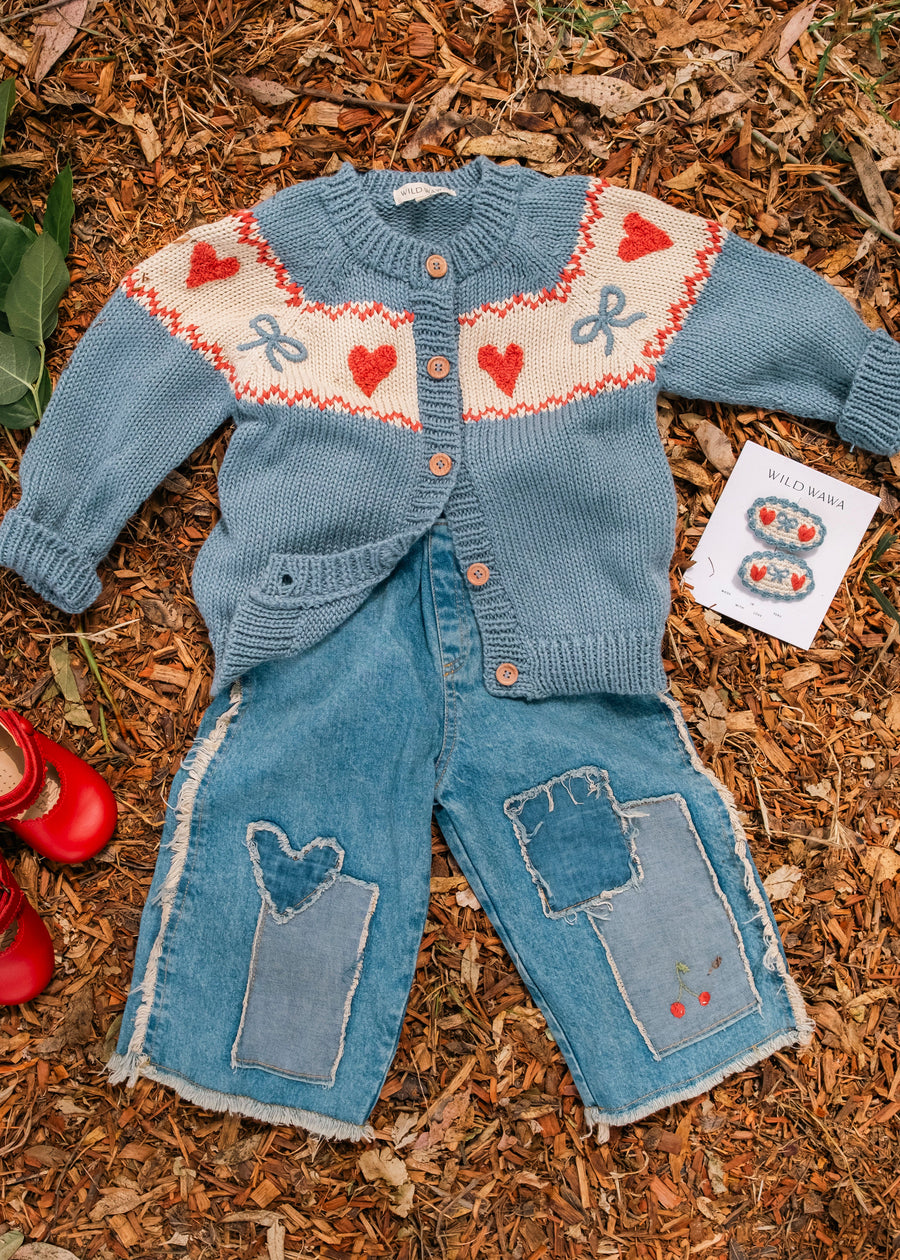 Children's blue knitted cardigan and jeans with heart and bow pattern on a wooden surface.