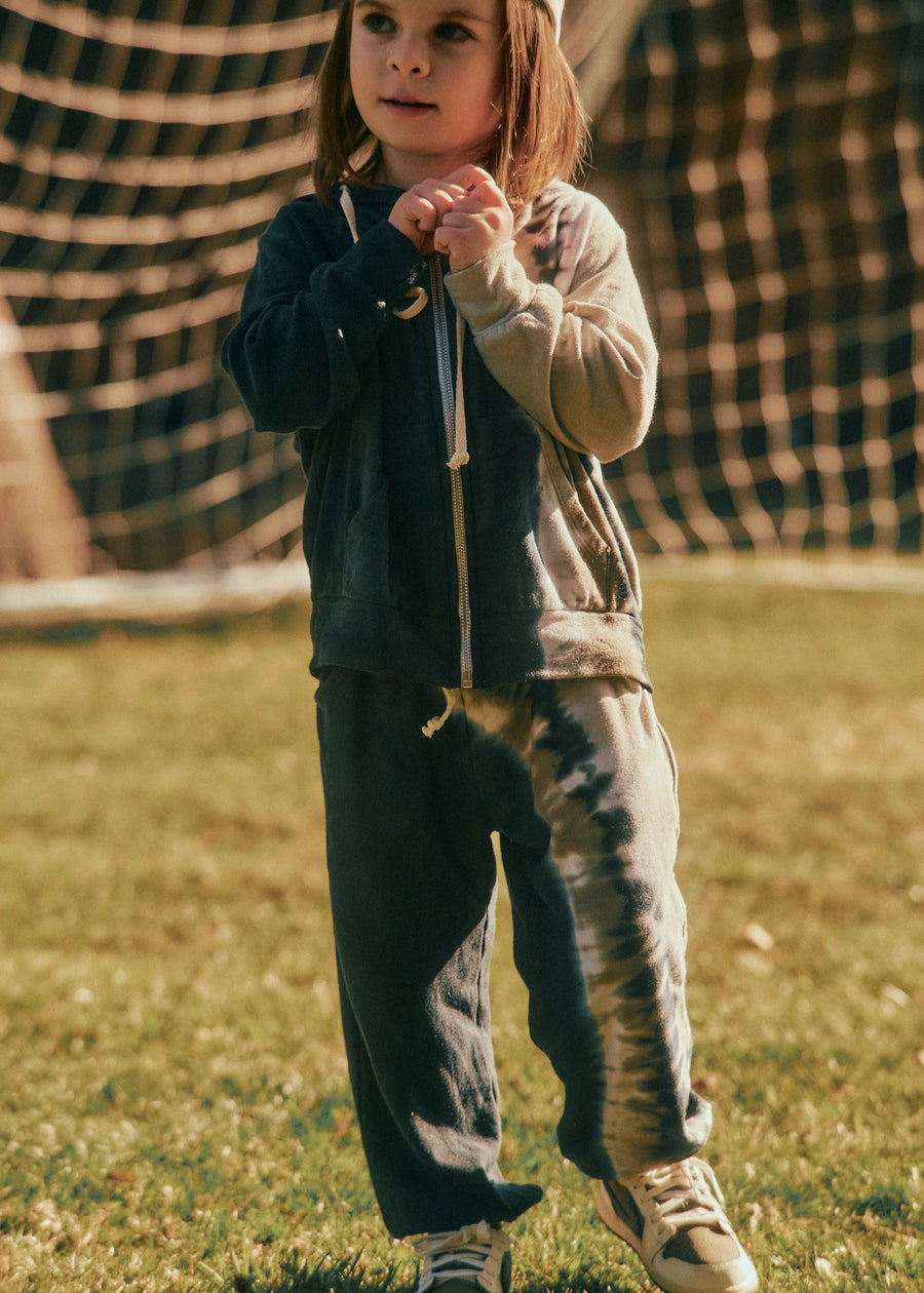 Child standing on grass wearing a dark jacket and light pants with a checkered tent in the background
