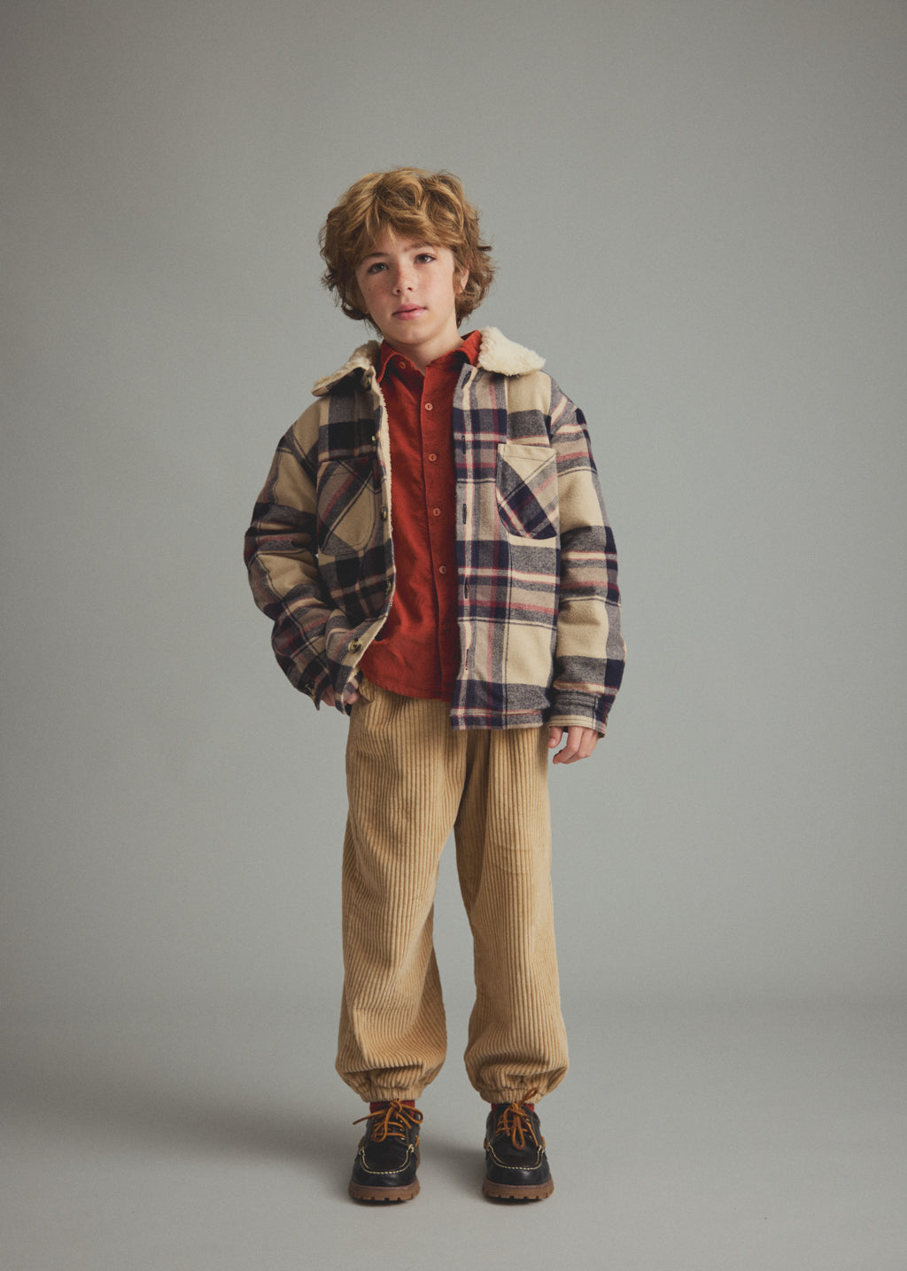 A young boy stands against a solid gray studio background wearing a beige and navy plaid sherpa-lined jacket over a rust red shirt, tan corduroy pants, and dark brown shoes.