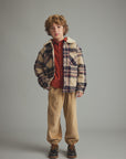 A young boy stands against a solid gray studio background wearing a beige and navy plaid sherpa-lined jacket over a rust red shirt, tan corduroy pants, and dark brown shoes.