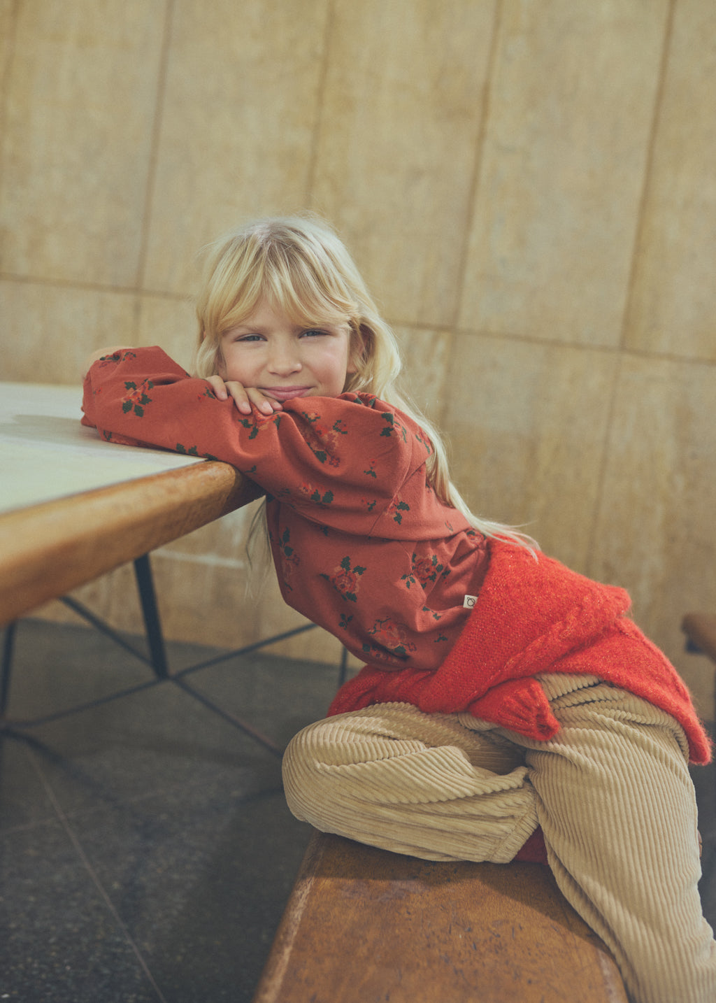 A young blonde girl sitting on a wooden bench in a cafe, wearing a rust-red patterned long-sleeve top and beige corduroy pants.
