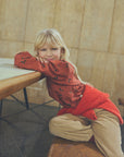A young blonde girl sitting on a wooden bench in a cafe, wearing a rust-red patterned long-sleeve top and beige corduroy pants.