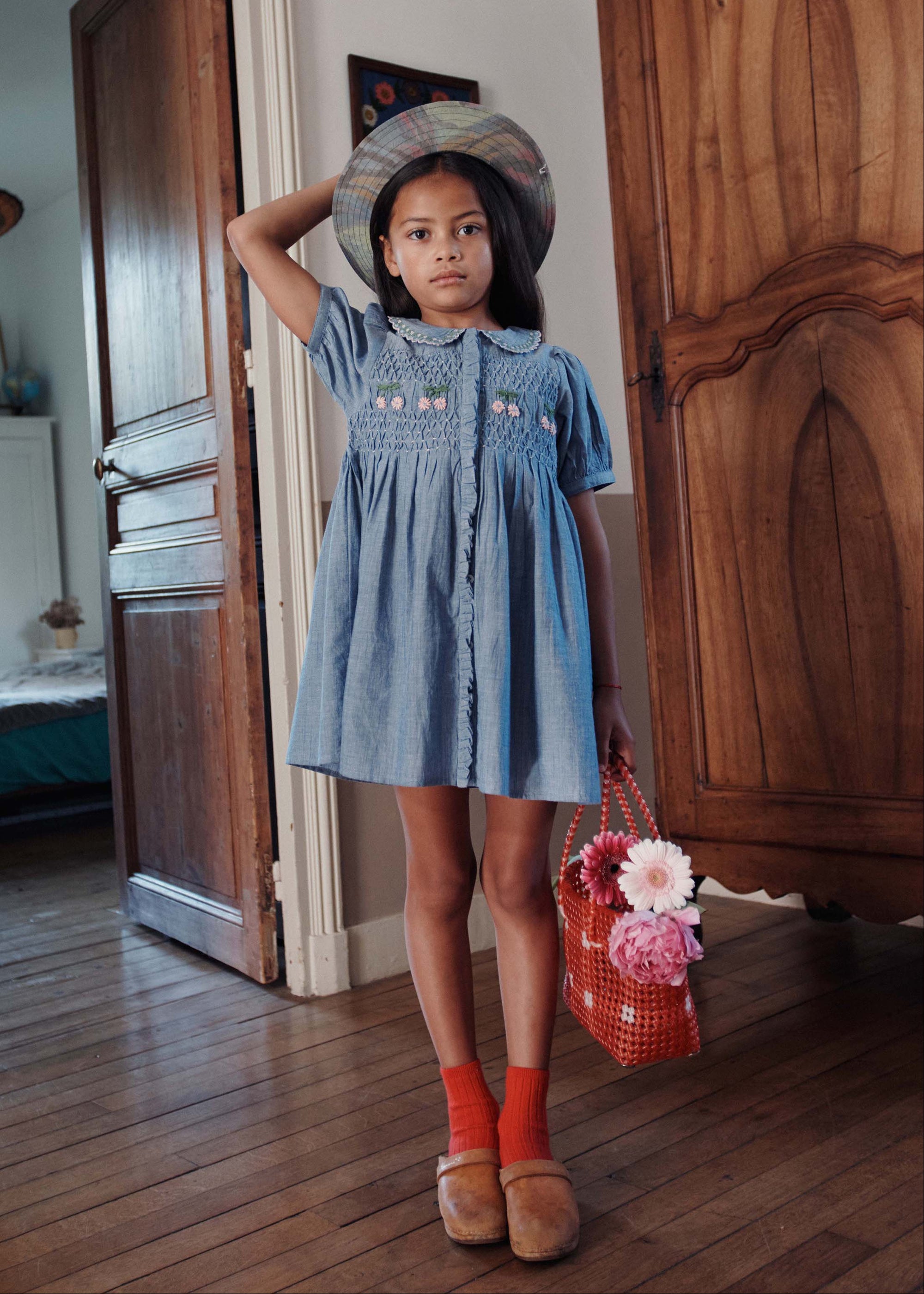 Young girl in a denim dress standing in a room with wooden furniture.