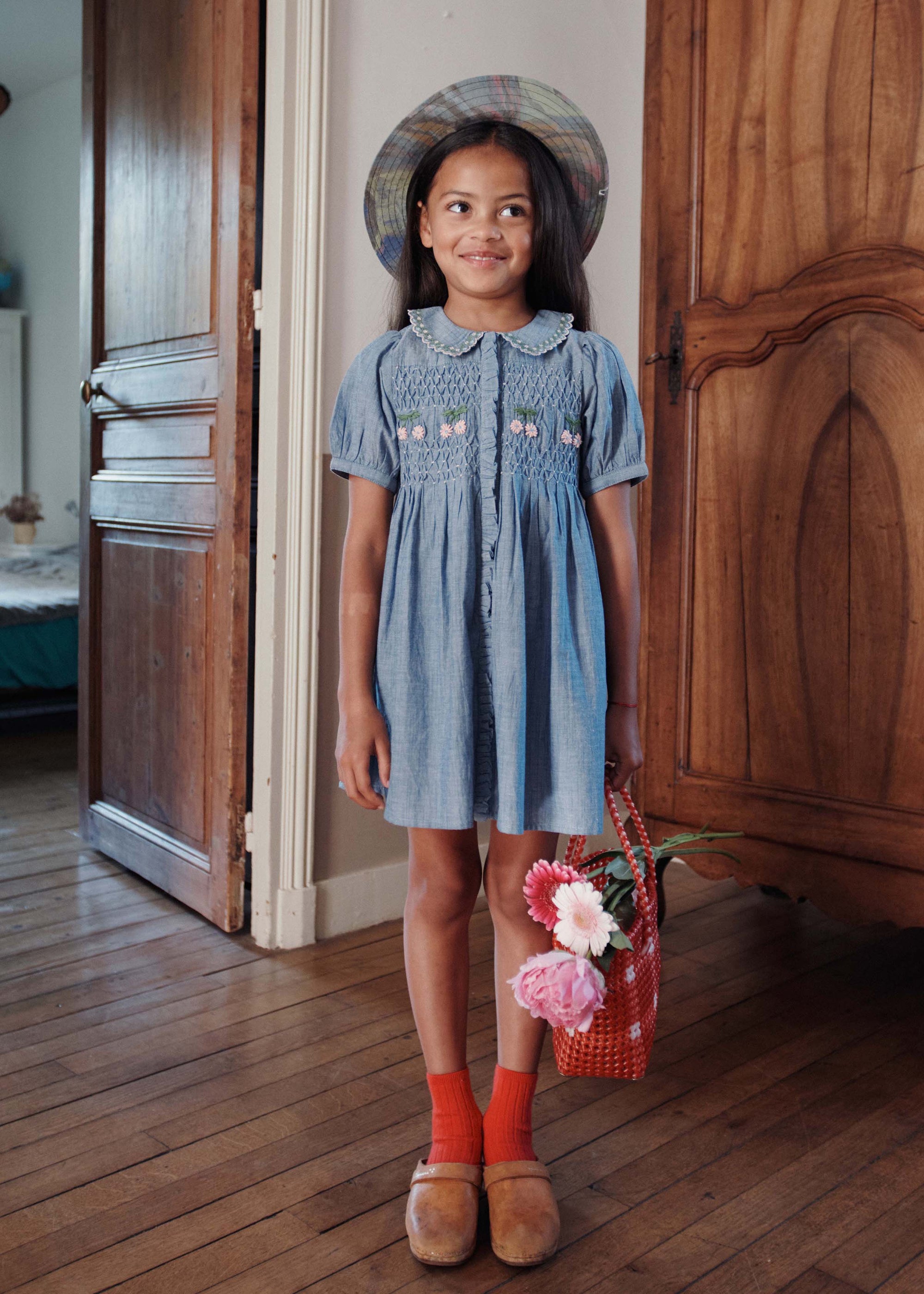 Young girl in a blue dress standing in a room with wooden floors and doors.