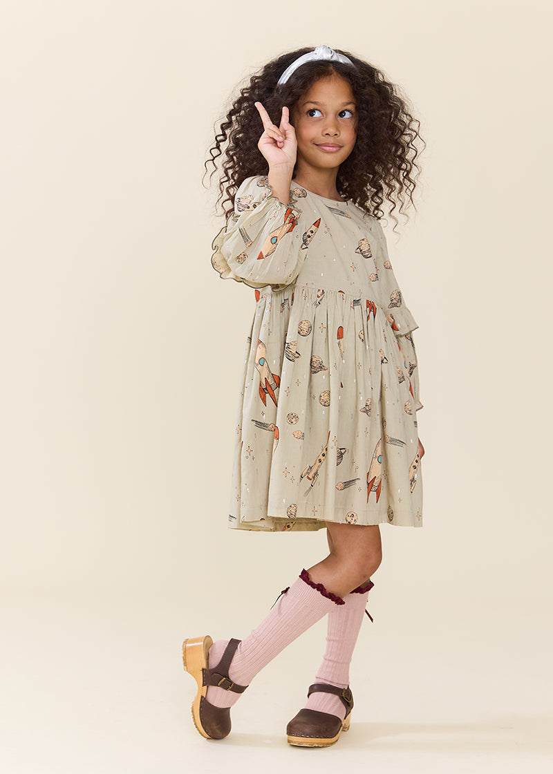 A young girl with curly dark hair wearing a beige tiered dress with a whimsical print, pink knee-high socks, and brown shoes, posing against a solid cream background.