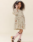A young girl with curly dark hair wearing a beige tiered dress with a whimsical print, pink knee-high socks, and brown shoes, posing against a solid cream background.