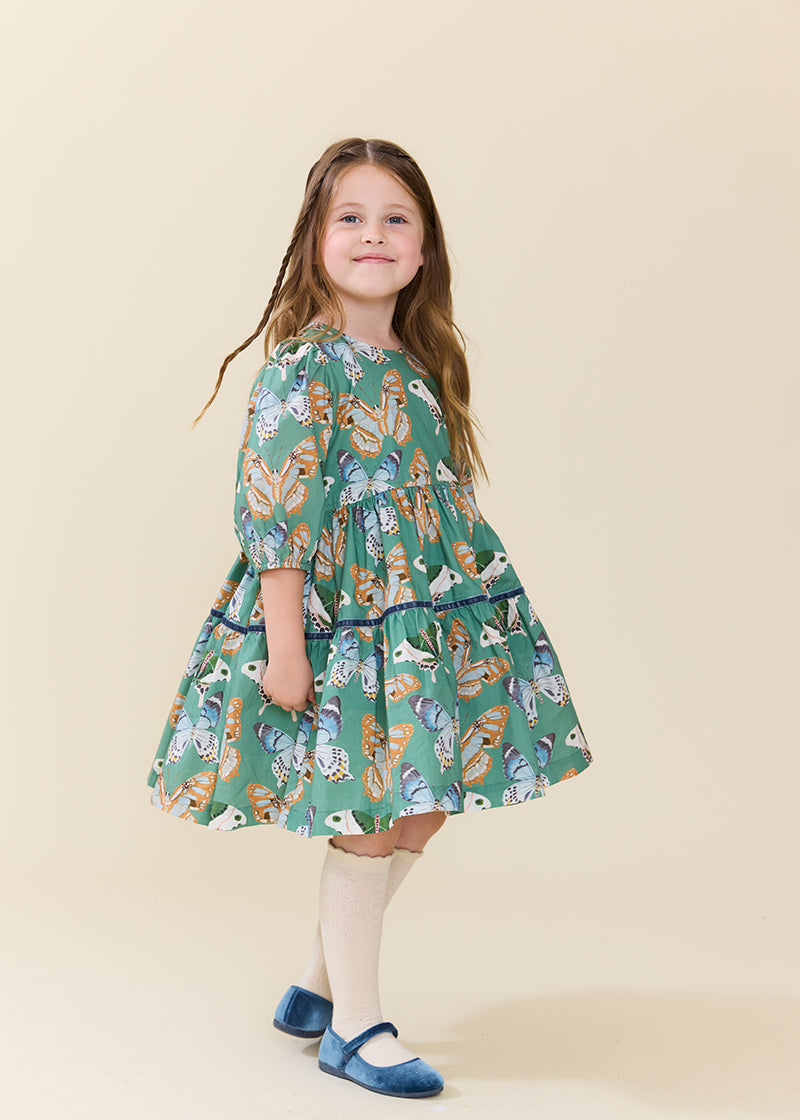 A young girl with long brown hair stands in a studio wearing a green tiered dress with a colorful butterfly and bird print, paired with cream tights and blue shoes, against a solid beige background.
