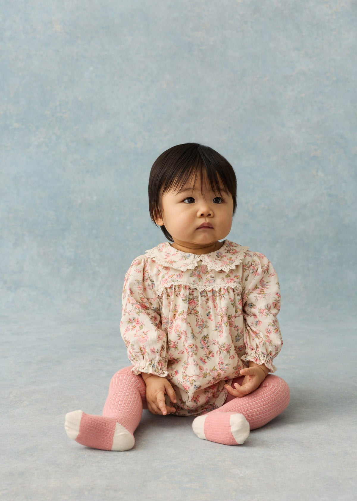 A baby girl sitting on a textured light blue studio background, wearing a long-sleeve cream and pink floral romper with a ruffled collar, pink ribbed tights, and pink-and-white socks.