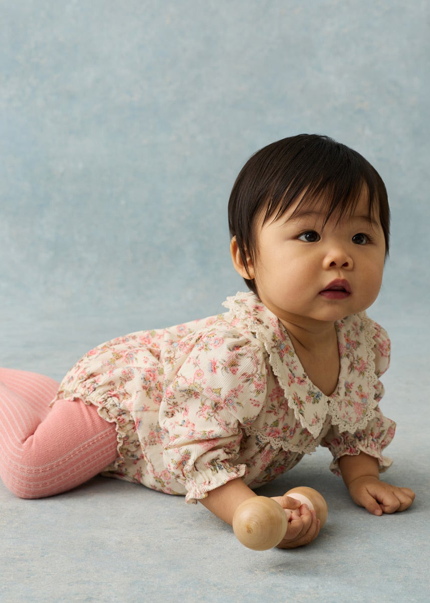 A baby girl lying on her stomach, wearing a light pink and white floral long-sleeve romper with pink ribbed tights, set against a textured light blue-gray studio backdrop.