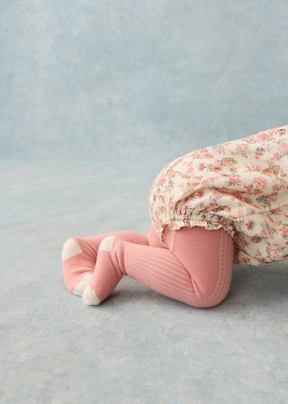 A close-up of a baby's legs wearing pink ribbed tights with white toe caps and a floral smocked diaper cover, kneeling on a textured light blue studio floor.