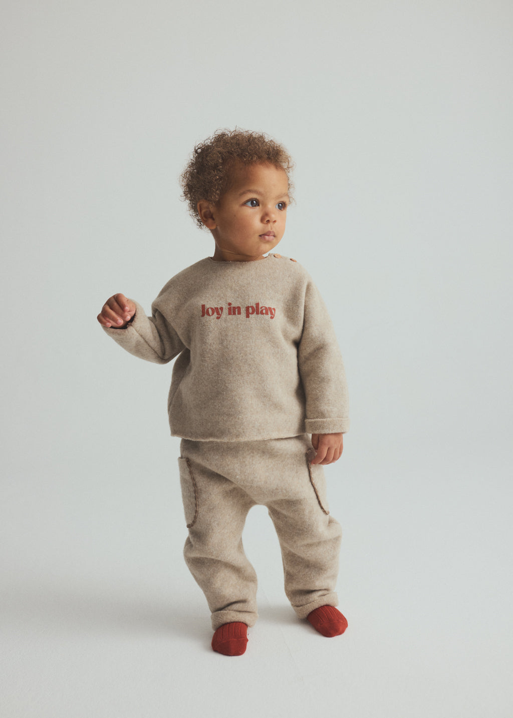 A toddler standing against a light gray studio background, wearing a beige knit two-piece outfit with red text reading 'joy in play' on the chest and red socks.