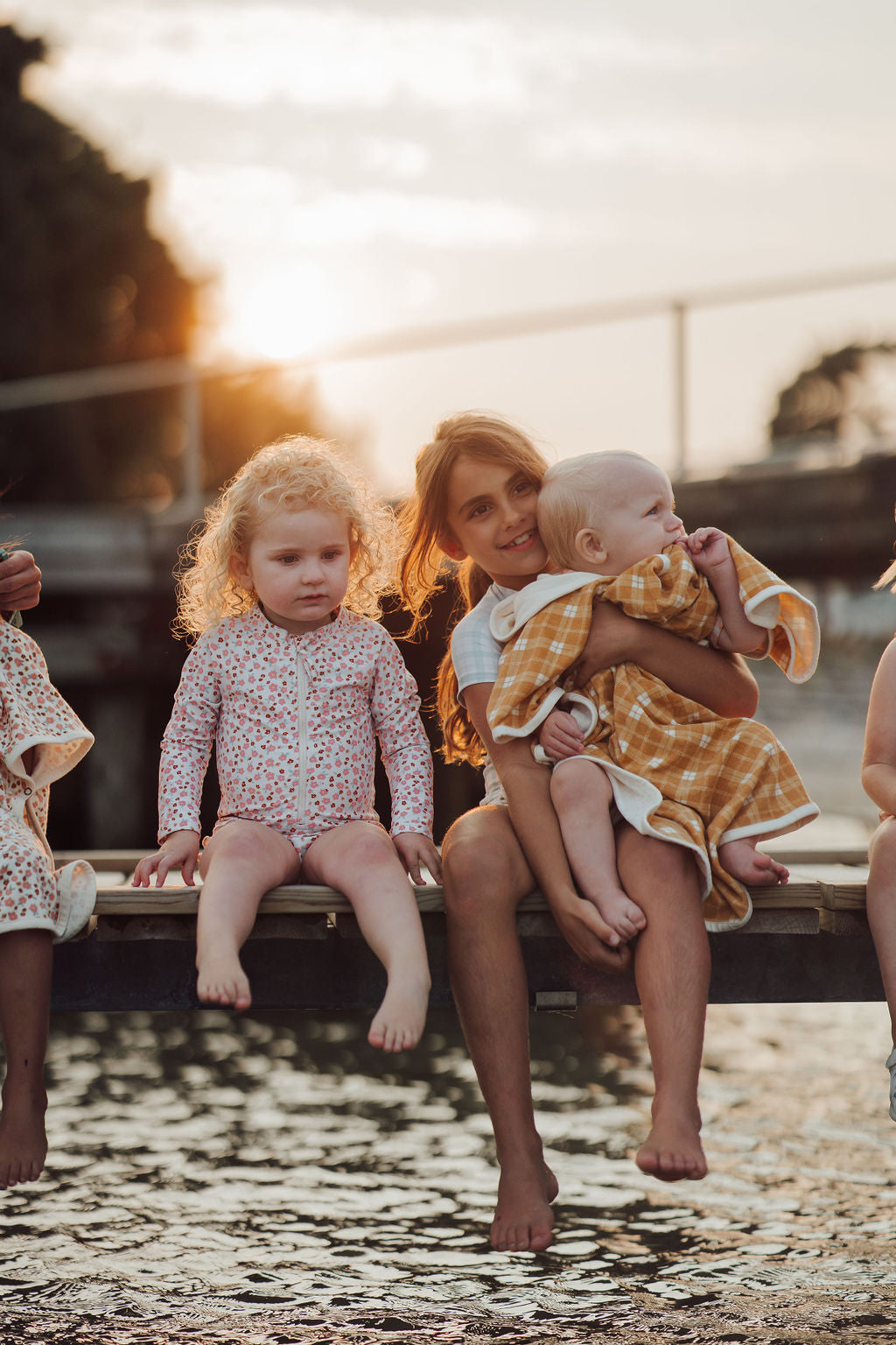 Woman holding a baby and sitting with two other children by a body of water at sunset.