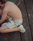 Child sitting on a wooden dock wearing checkered shorts.
