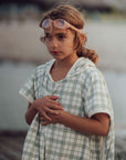 Young girl wearing goggles and a checkered dress by a body of water