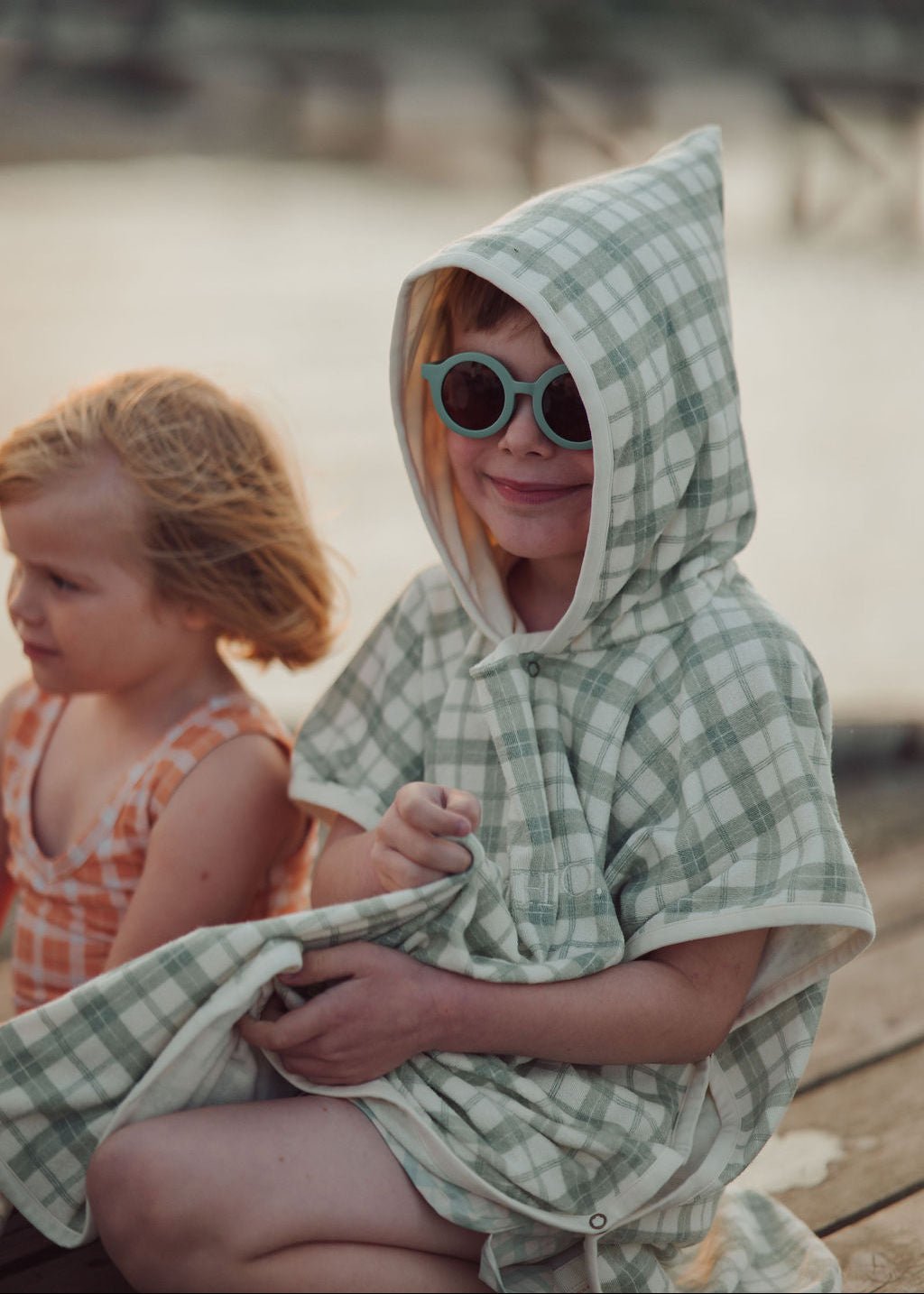 Child wearing a green checkered poncho sitting by a body of water with another child in the background.