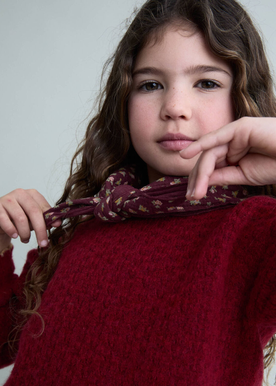 Young girl wearing a red sweater with a patterned scarf, posing against a plain background.