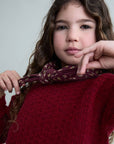 Young girl wearing a red sweater with a patterned scarf, posing against a plain background.
