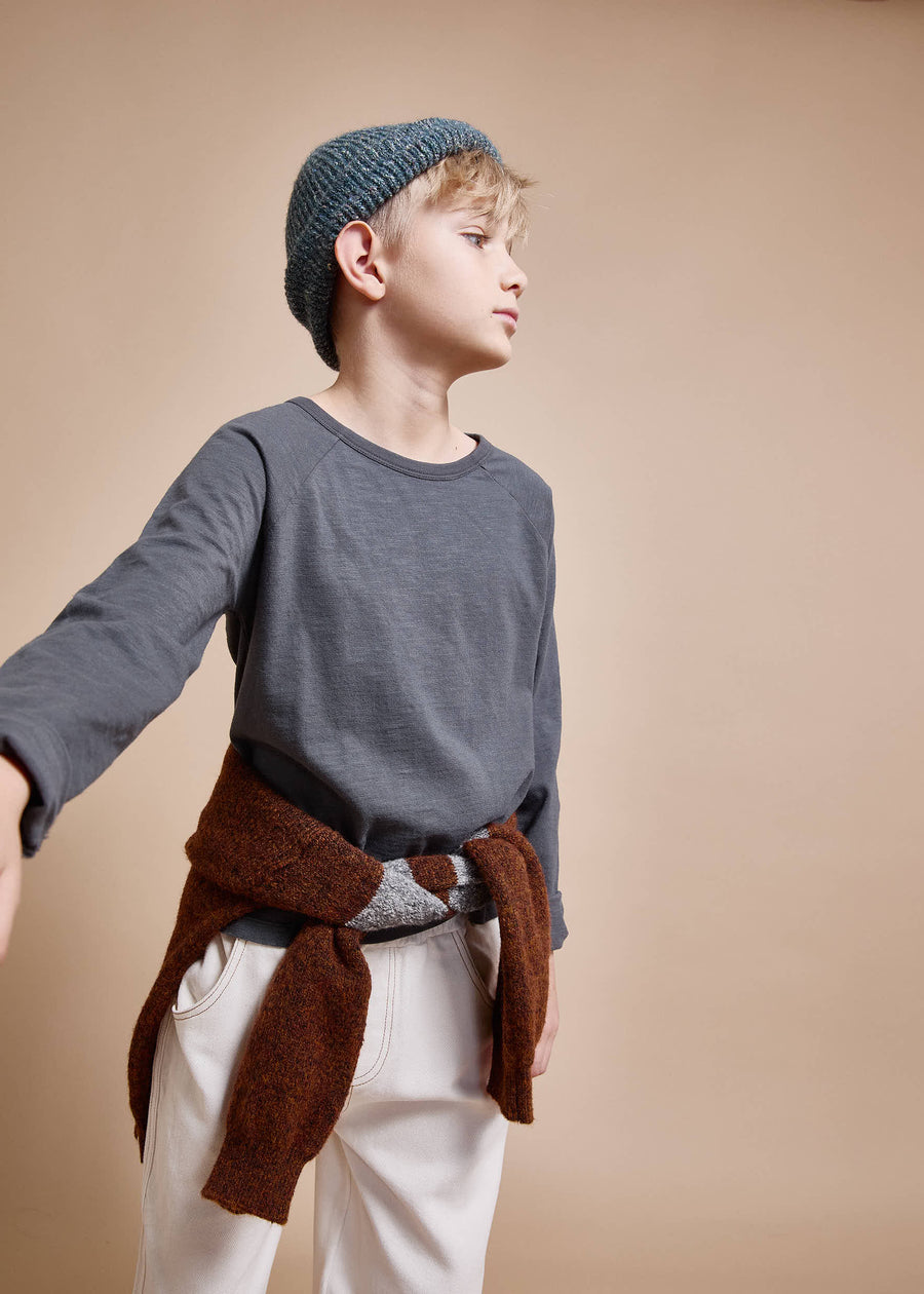Child wearing a gray sweater, white pants, and a blue knitted hat against a beige background