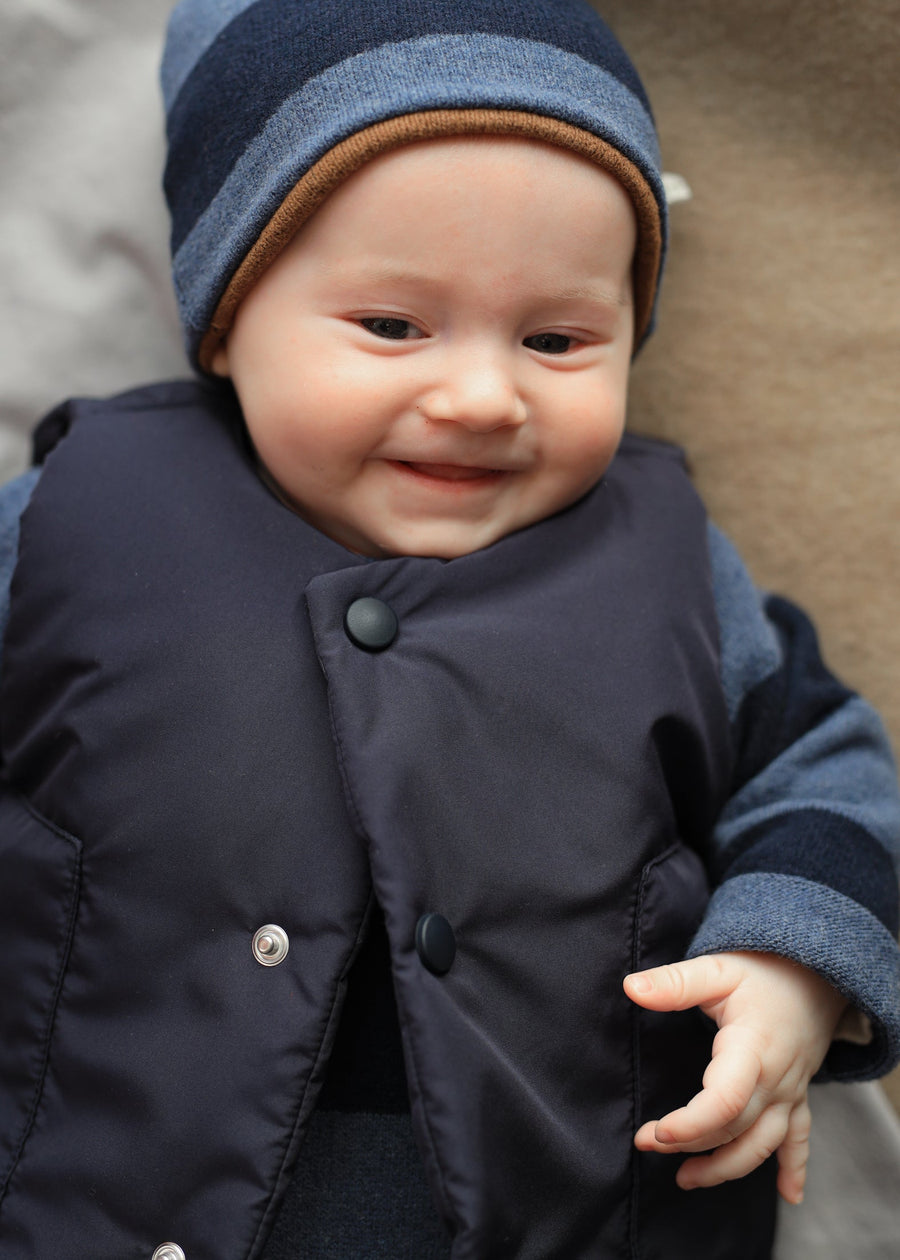 Baby wearing a navy coat and striped hat with a brown interior, smiling.