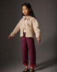 Young girl wearing a cream sweater with a bow and patterned burgundy pants on a gray background