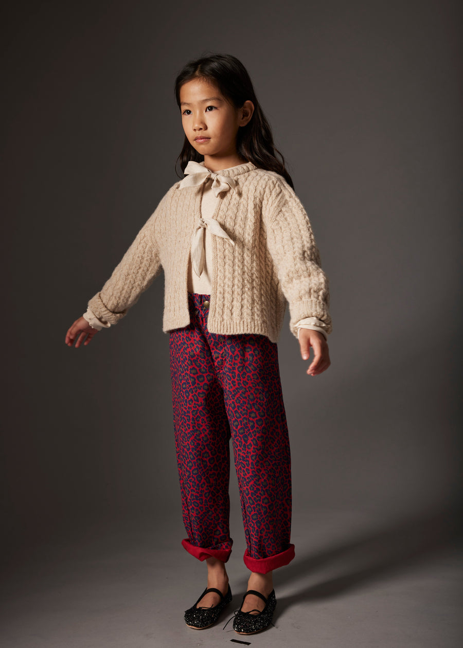 Young girl wearing a cream sweater with a bow and patterned burgundy pants on a gray background