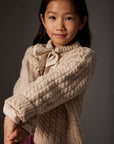 Young girl wearing a beige cable knit sweater with a bow tie against a gray background