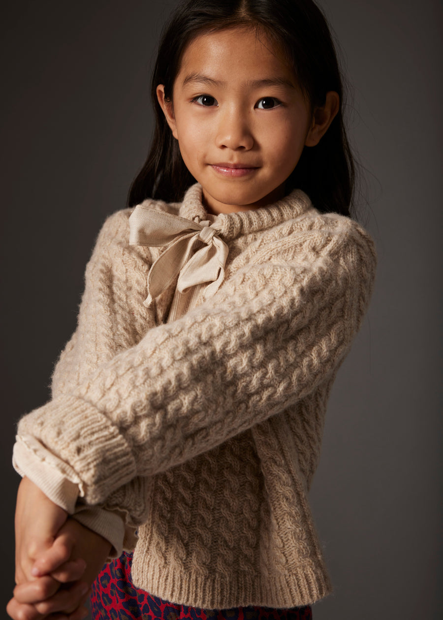 Young girl wearing a beige cable knit sweater with a bow tie against a gray background