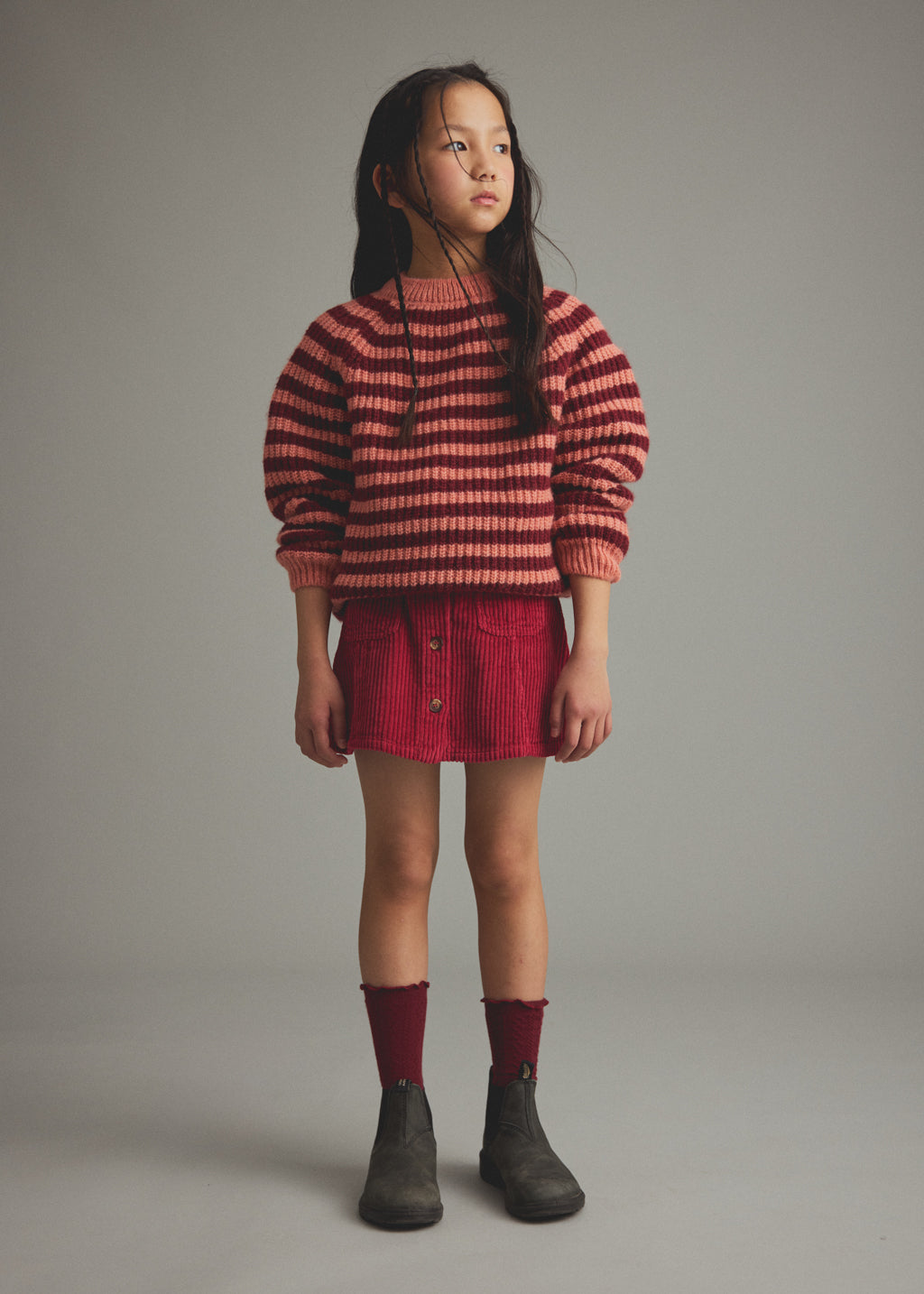 A young girl stands against a neutral gray studio background wearing a red and pink striped knit sweater, a red corduroy mini skirt, dark red socks, and dark gray boots.