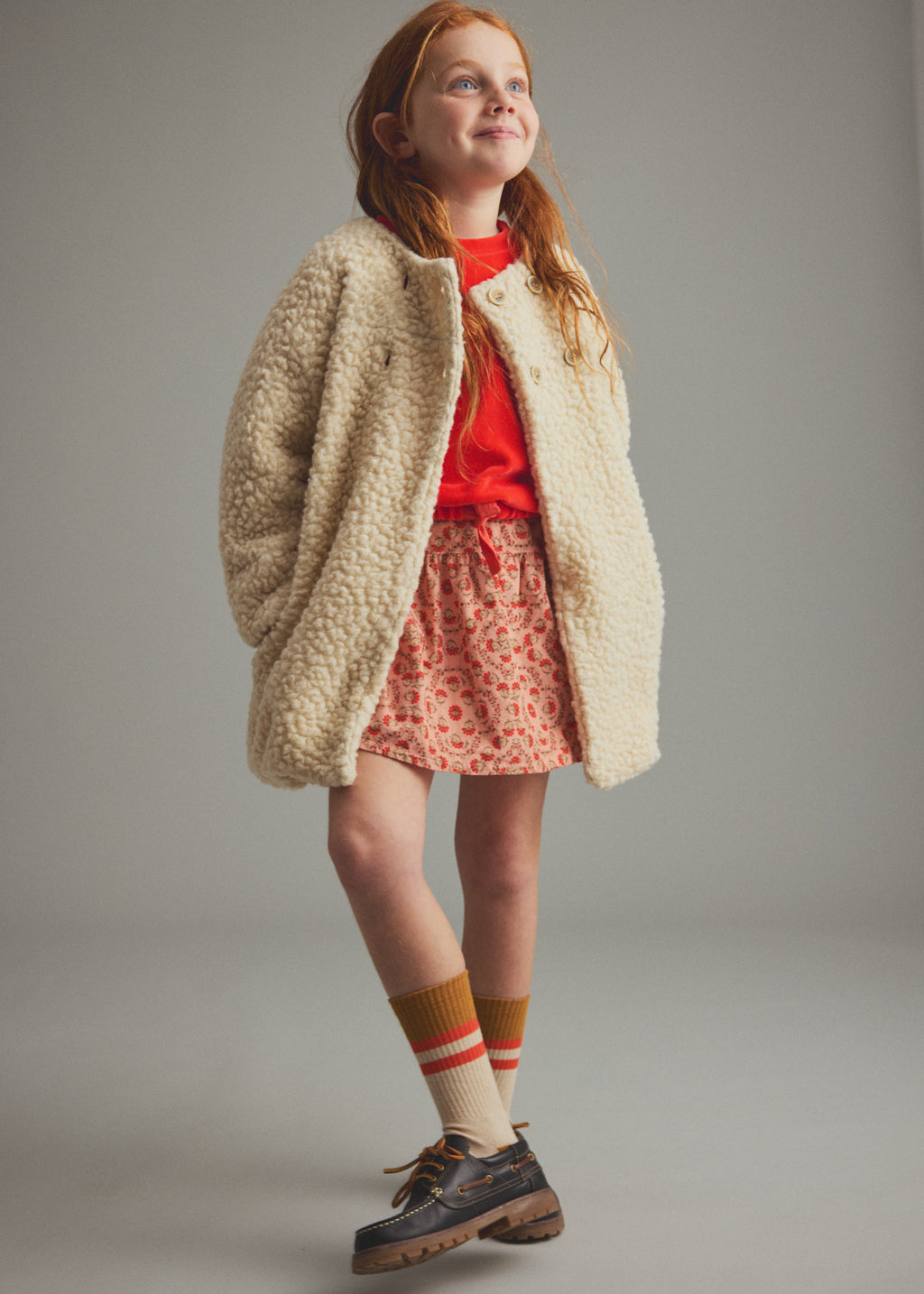 A young girl with red hair stands against a neutral gray studio background wearing a cream sherpa fleece jacket over a bright red top, a red and white patterned skirt, striped socks, and dark brown shoes.